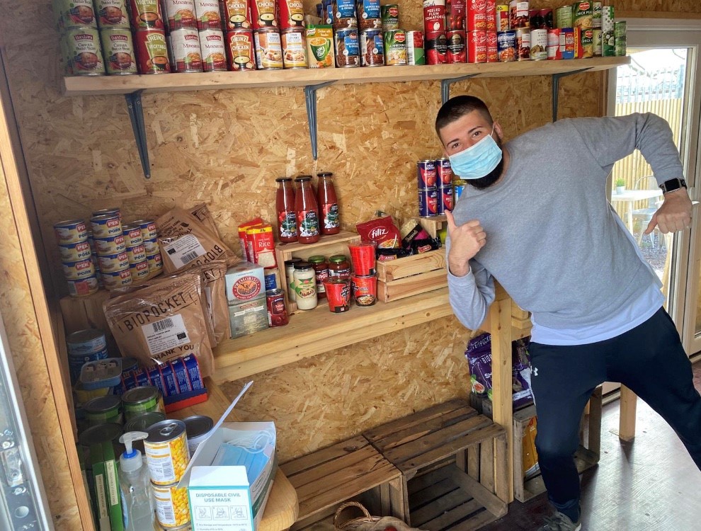 A man wearing a face mask pulls a thumbs up pose in front of wooden shelves of food